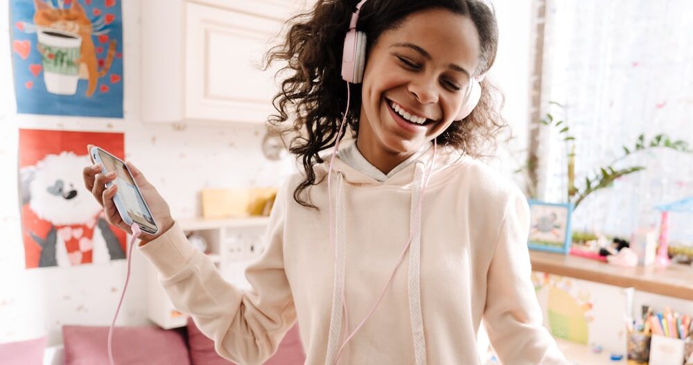Happy teen in headphones listening to music and dancing in her cozy room at home