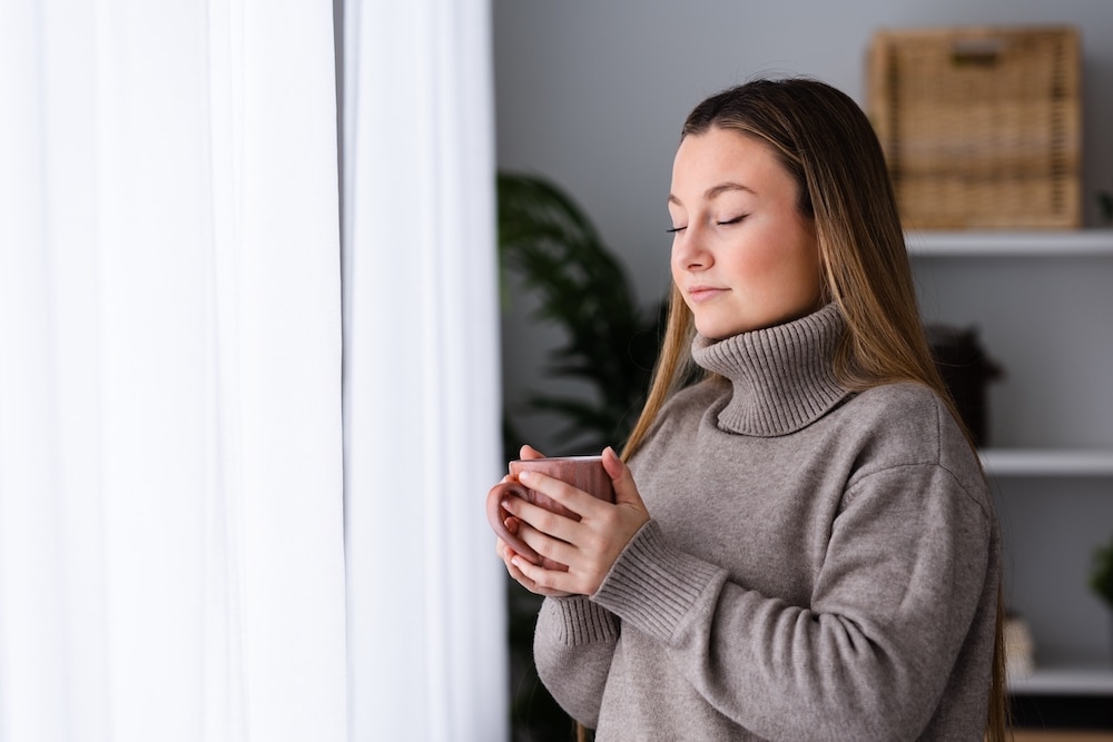 Winter clothing young woman breathing deep beside window while holding cup of hot drink as tea or coffee.